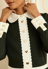 Černé šaty s bílým vroubkovaným límečkem, zdobené zlatými knoflíky ve tvaru srdce podél přední části a manžet. Hladká textura látky.
