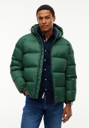 Giovane uomo che indossa una giacca imbottita verde sopra una camicia blu scuro con bottoni e jeans blu, in piedi davanti a uno sfondo bianco.