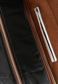 Barna gyapjú és fekete nylon anyagú zipzáras dzseki belseje, fehér csíkos zsebbe varrt zipzárral és részletes varrással.