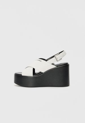White platform sandals with a black base, featuring a crossed strap design and adjustable ankle buckle. Smooth texture, modern shape.