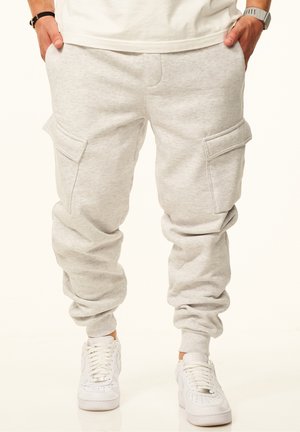 Man wearing light gray cargo sweatpants with elastic cuffs and white sneakers, hands in pockets, standing against plain background.