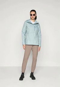 Gaiši zila ūdensnecaurlaidīga jaka ar jostu krūšu kabatā, apvienota ar beigtu biksēm un melniem apaviem. Modelis valkā saulesbrilles.