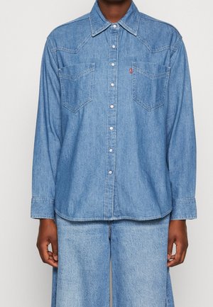 Personne portant une chemise en denim bleu clair à boutons avec poches poitrine et un pantalon en denim assorti sur un fond uni.