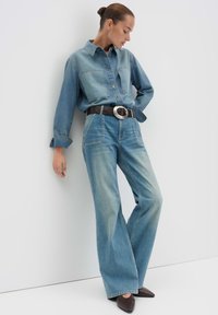 Chemise en denim à manches longues, avec deux poches poitrine ; associée à un jean large bleu clair. Ceinture noire avec une boucle circulaire en argent. Chaussures pointues marron.
