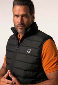 Schwarze Daunenweste mit hohem Kragen, Reißverschluss und Logo auf der Brust, über einem orangefarbenen Poloshirt getragen. Glatte Textur und gestepptes Design.