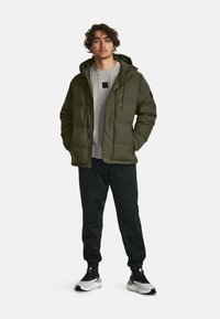 Groene pufferjack met capuchon, gecombineerd met een lichtgrijs sweater en zwarte broek, met witte en zwarte sport schoenen.