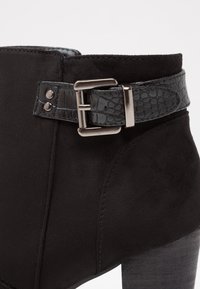Bottines noires avec une sangle à boucle en faux cuir texturé, tige en daim lisse et talon empilé. Présente des accents en métal argenté.