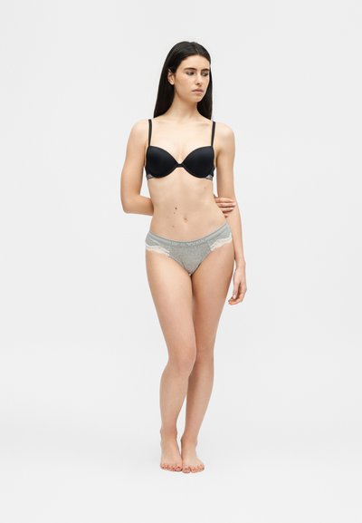 Γυναίκα που στέκεται ξυπόλυτη, φοράει μαύρο σουτιέν και γκρι εσώρουχο με δαντέλα, ποζάροντας με το ένα χέρι πίσω από την πλάτη της μπροστά σε απλό φόντο.