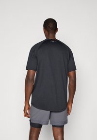 T-shirt de sport noir à manches courtes, en tissu lisse. Offre un col rond et un petit logo à l'arrière. Associé à un short gris.