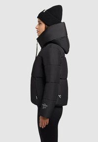 Sort pufferjakke med høj krave, lynlåslommer og subtil branding. Glat tekstur med en tætsiddende silhuet. Sort strikket beanie.