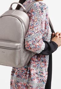 Hellgrauer Leder-Rucksack mit runder Form, vorderer Reißverschlusstasche, verstellbaren Trägern und sichtbarem Markenlogo. Der Strickpullover hat eine mehrfarbige Textur.