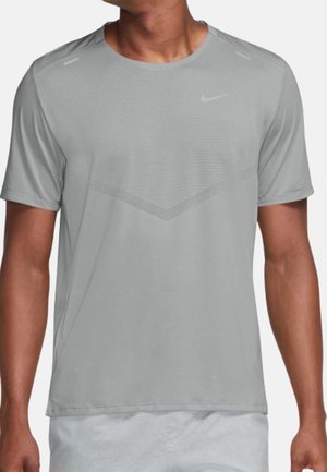 Mann trägt hellgraues Nike Sportshirt mit kurzen Ärmeln und helle Shorts, steht vor einfachem Hintergrund.