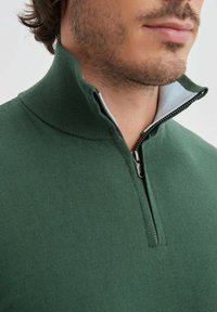 Pull over vert à fermeture éclair quart de longueur, fabriqué en tissu doux. Doté d'un col montant avec une doublure intérieure contrastante et d'une fermeture éclair en métal.