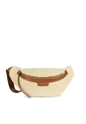 Flødefarvet shearling bumbag med brune læderkanter, lynlås og justerbar brun rem, med mærket "Lancaster Paris" foran.