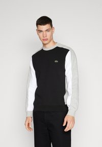 Felpa nera con maniche grigie e bianche, caratterizzata da un piccolo logo verde Lacoste sul petto. Realizzata in morbido tessuto misto cotone.