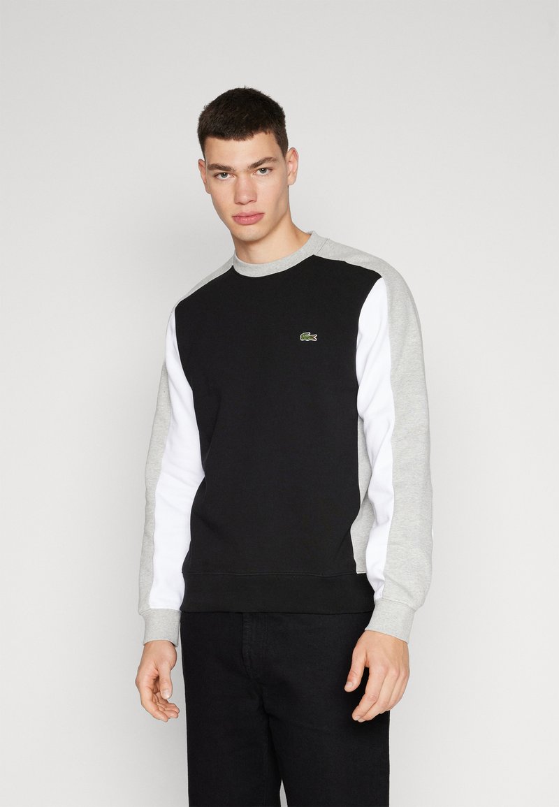 Felpa nera con maniche grigie e bianche, caratterizzata da un piccolo logo verde Lacoste sul petto. Realizzata in morbido tessuto misto cotone.