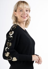 Pulover negru cu mâneci lungi, decorat cu modele de inimi din paiete aurii. Textură moale, manșete cu nervuri și un design clasic cu gât rotund.