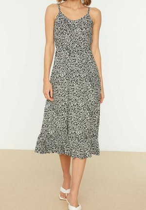 Femme portant une robe midi noire et blanche à fleurs sans manches avec un ourlet à volants, associée à des talons blancs ouverts.