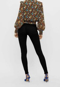 Blommig blus med lång ärm och rynkade manschetter på svart bakgrund, kombinerad med svarta tajta jeans och blå klackar med remmar.