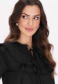 Sort bluse med lagdelt flæsedesign med kant, nøglehulslukning bundet med et bånd og lange ærmer. Strukturstof.