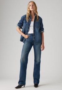 Denim jacket met twee voorzakken, lichtblauw. Witte tanktop eronder. Donkerblauwe flared jeans. Zwarte hakken mules. Neutrale achtergrond.