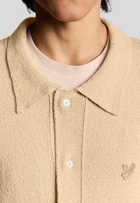 Beige strukturmönstrad polo skjorta med krage, prydd med tre knappar och en broderad logotyp på bröstet. Lagd över en ljusrosa t-shirt.