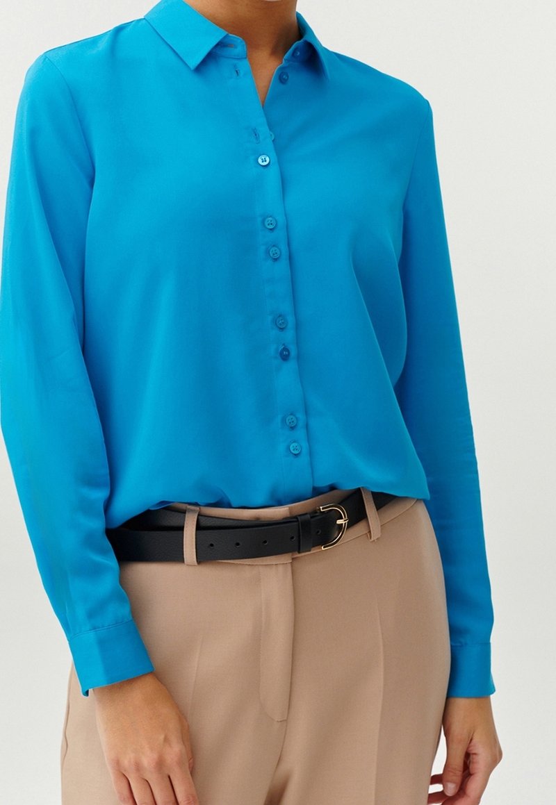 Femme portant une chemise bleu vif à manches longues boutonnée, rentrée dans un pantalon beige avec une ceinture noire à boucle dorée.