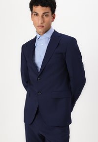Navy suit featuring a two-button closure, notch lapels, and side pockets. Paired with a light blue dress shirt underneath.