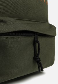 Zielony plecak z płótna z zamknięciem na górze i przednią kieszenią z czarnym zamkiem błyskawicznym oraz szczegółami sznurka.