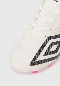 Botas de fútbol en blanco con una parte superior texturizada, diseño geométrico negro y acentos rosas en la suela. Presenta un sistema de cordones tradicional.