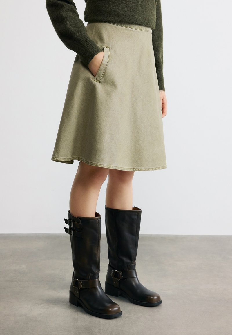 Γυναίκα που φοράει ελιά-πράσινη φούστα Α-γραμμή μέχρι το γόνατο με πλαϊνές τσέπες, σκούρο πράσινο πουλόβερ και μαύρες δερμάτινες μπότες μέχρι το γόνατο με αγκράφες.