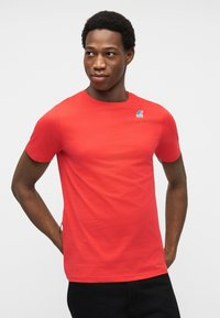 Άνδρας που φοράει κόκκινη κοντομάνικη μπλούζα με μικρό μπλε και κόκκινο λογότυπο στο αριστερό στήθος, στέκεται με τα χέρια πίσω από την πλάτη.