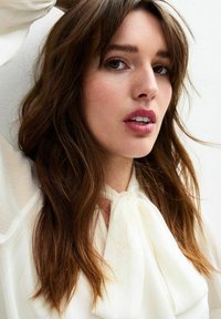 Close-up of a woman with long, wavy brown hair, wearing a white blouse with a tie collar, featuring soft, textured fabric and natural makeup.