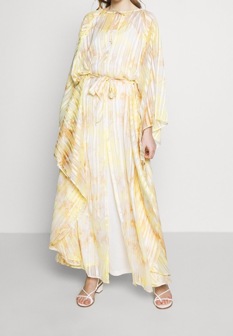 Femme portant une robe maxi plissée fluide jaune pastel et blanche, à manches longues, avec une ceinture à la taille et des sandales blanches à brides.