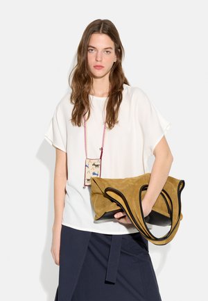 Młoda kobieta o długich brązowych włosach nosi białą bluzkę, granatową spódnicę, trzyma dużą jasno-brązową zamszową torbę z czarną lamówką oraz małą wzorzystą saszetkę na naszyjniki.