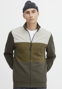 Giacca con zip caratterizzata da pannelli verdi e beige, realizzata in un tessuto morbido con finitura texturizzata. Collo alto e dettagli con zip frontale.