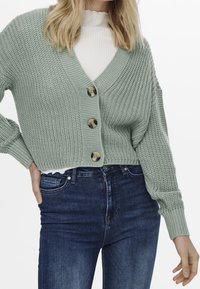 Kvinna iklädd en ljusgrön grovstickad kort cardigan med stora knappar över en vit ribbad polotröja och mörkblå jeans.