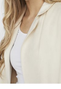 Vaaleanbeige takki, jossa on sileä pinta, räätälöity kaulus ja avoin edusta, kerrostettuna valkoisen paidan päälle.