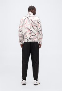 Άνδρας που στέκεται με την πλάτη γυρισμένη, φορώντας λευκό μπουφάν με μαύρες και κόκκινες αφηρημένες γραμμές, μαύρο παντελόνι και λευκά αθλητικά παπούτσια σε λευκό φόντο.