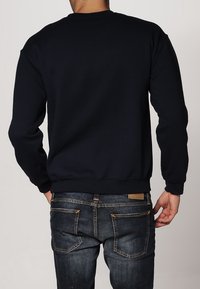 Uomo che indossa una felpa nera a maniche lunghe e jeans blu scuro, girato di spalle con le mani vicino alle tasche contro uno sfondo neutro.
