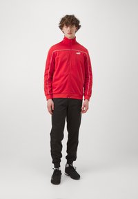 Červená bunda na zips s bielymi akcentmi, v kombinácii s čiernymi teplákmi a čiernymi teniskami. Bunda má vysoký golier a logo na hrudi.