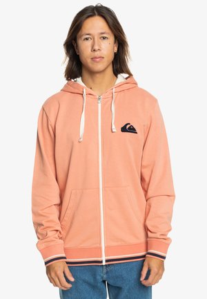 Peach zip-up hoodie in soft fabric with a white lining, featuring a navy logo, ribbed cuffs, and striped hem. Two front pockets.