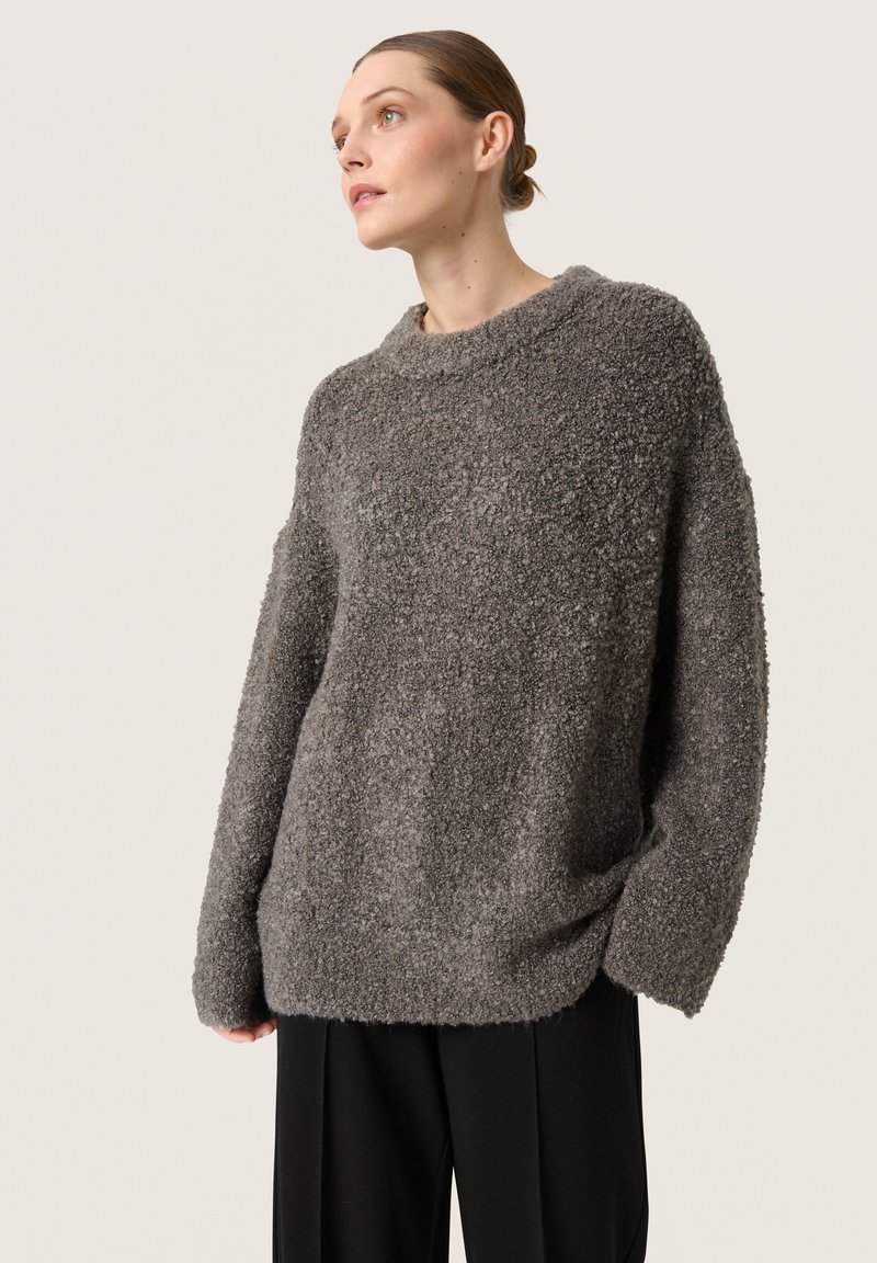 Oversized grå sweatshirt lavet af et tekstureret, fluffy stof, med bred halsudskæring og løstsiddende ærmer, parret med sorte bukser.