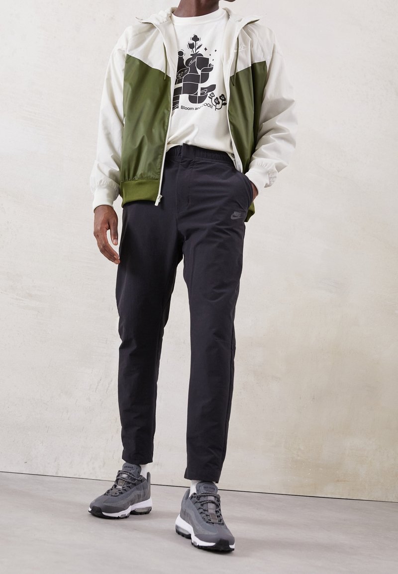 Giacca sportiva verde oliva e bianca, t-shirt grafica con design nero, pantaloni neri e scarpe da ginnastica grigie con suola bianca. Vestibilità slim, stile casual.