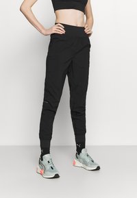 Schwarze Sporthose mit elastischem Bund, zulaufenden Beinen und seitlichen Akzenten. Kombiniert mit grauen und pinken Sneakers sowie schwarzen Knöchelsocken.