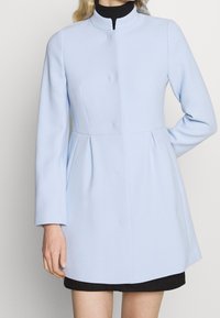 Donna che indossa un cappotto aderente blu chiaro con maniche lunghe sopra un vestito nero, in piedi con un braccio piegato dietro la schiena.