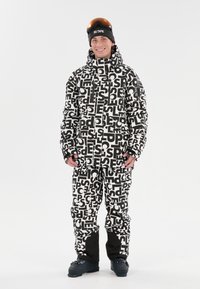 Lumityyli lumilautailuun, jossa musta-valkoinen graafinen printti takissa ja housuissa, korkea kaulus, vetoketjut ja mustat saapasdetaljit.