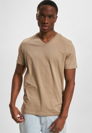 Ung mann med kort hår iført en beige V-hals T-skjorte og lyseblå jeans, stående mot en ensfarget grå bakgrunn.
