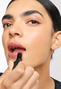 Se aplica lápiz labial mate en los labios. El tubo tiene un diseño rectangular negro. La piel tiene un tono natural y suave con ligeros toques de maquillaje.
