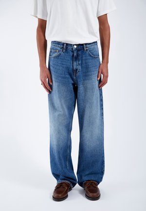 Light blue faded denim jeans with a relaxed fit, featuring five pockets and a button fly. Worn with a white short-sleeve t-shirt and brown shoes.